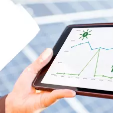 Uma mão segurando um tablet, que contém a imagem de um gráfico de monitoramento de geração de energia fotovoltaica